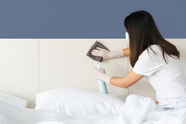 Фото Молодая азиатка в медицинской маске, руки в резиновой защитной перчатке с тканью из микроволокна и бутылкой с моющим средством, моющая поверхность в спальне дома