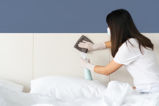 Young Asian woman wearing medical mask hands in rubber protective glove with micro fiber cloth and detergent spray bottle cleaning a surface in bedroom at home Closeup