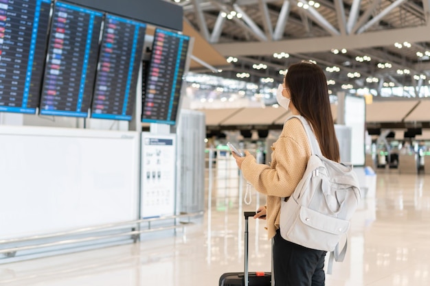 国際空港で医療用マスクを身に着けている若いアジア人女性が、コロナウイルスのパンデミックの新しい通常のライフスタイルの概念の間に彼女の飛行をチェックしているフライト情報ボードを見て