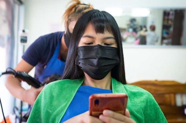 Giovane donna asiatica che indossa una maschera di protezione e usa lo smartphone mentre si asciuga i capelli con l'asciugacapelli dal parrucchiere. parrucchiere che asciuga i capelli al cliente al parrucchiere.