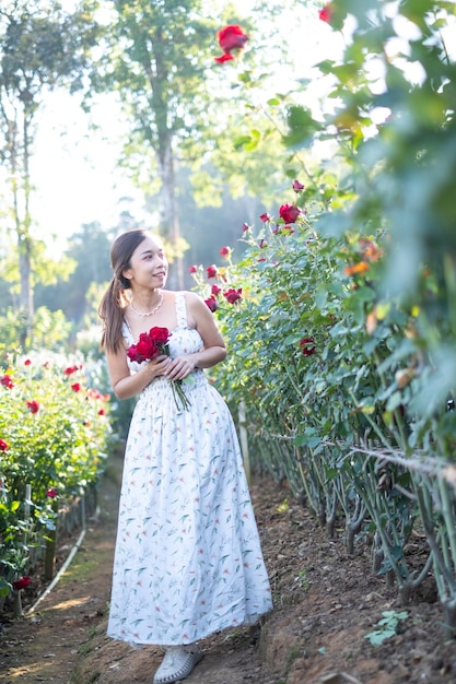 Young Asian woman wearing a white dress poses with a rose in rose garden