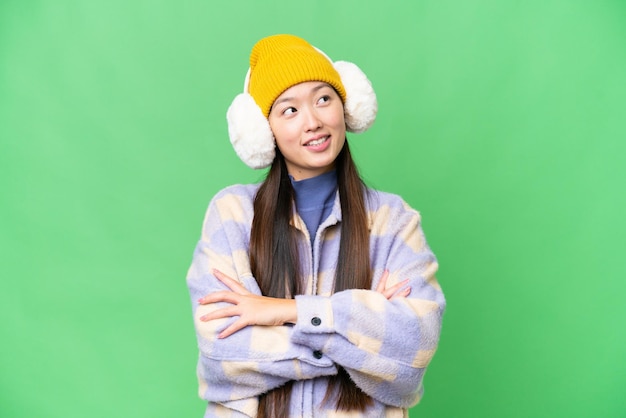 Young Asian woman wearing winter muffs over isolated chroma key background looking up while smiling