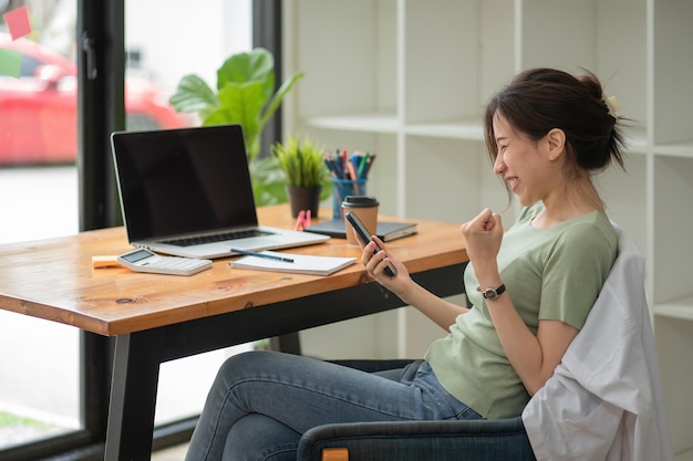 Young Asian woman winner celebrate victory triumph with a smartphone while sitting in modern office room