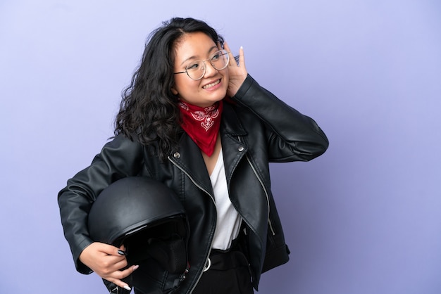 보라색 배경에 오토바이 헬멧을 쓴 젊은 아시아 여성이 귀에 손을 대고 무언가를 듣고 있다