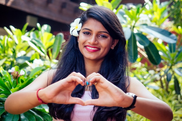 Young asian woman with black hair plumeria and fresh skin looking and smiling at the camera on park summer background hands in the shape of a heart valentine day