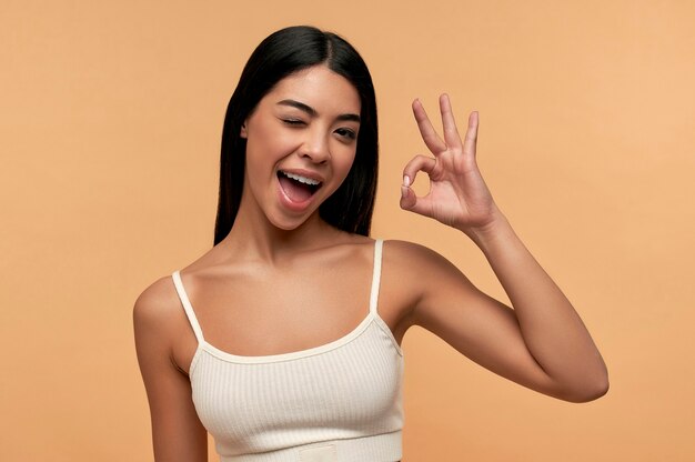 Young asian woman with clean healthy glowing skin in white top shows ok gesture isolated on beige