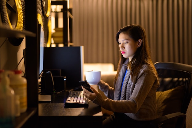 늦은 밤 집에서 일하는 동안 전화를 사용하여 커피와 함께 젊은 아시아 여자