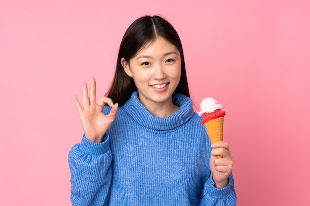 Young asian woman with a cornet ice cream isolated on pink wall showing ok sign with fingers