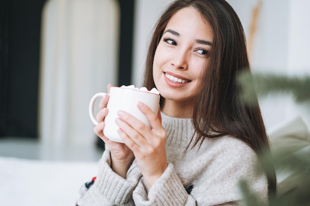 Young asian woman with cup of cocoa with marshmallow in room with christmas tree at home