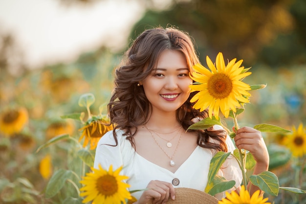 日没時のひまわり畑で巻き毛の若いアジアの女性。太陽の下で若い美しいアジアの女性の肖像画。夏。