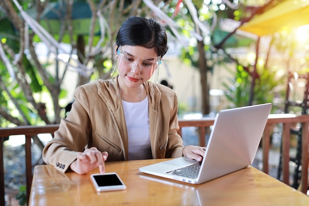 Giovane donna asiatica con visiera che si siede nella caffetteria e utilizza sullo smartphone e lavora al computer portatile. nuovo concetto di allontanamento normale e sociale