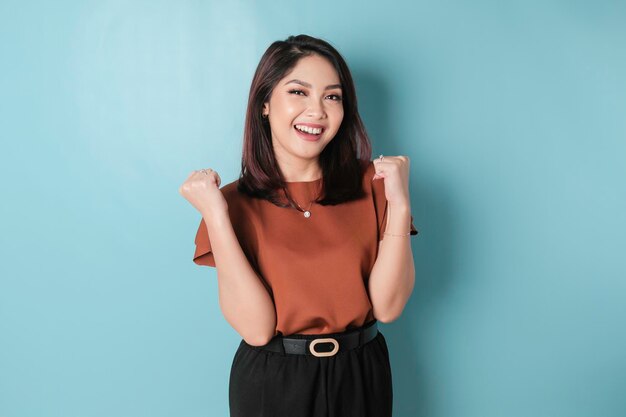 A young asian woman with a happy successful expression wearing\
brown shirt isolated by blue background