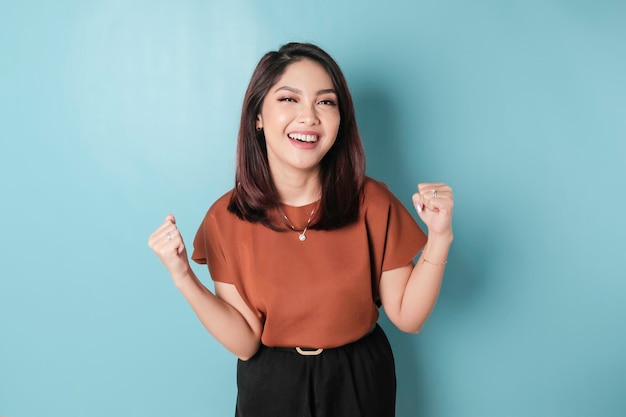 A young asian woman with a happy successful expression wearing\
brown shirt isolated by blue background