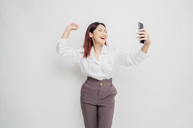 A young Asian woman with a happy successful expression wearing white shirt and holding smartphone isolated by white background