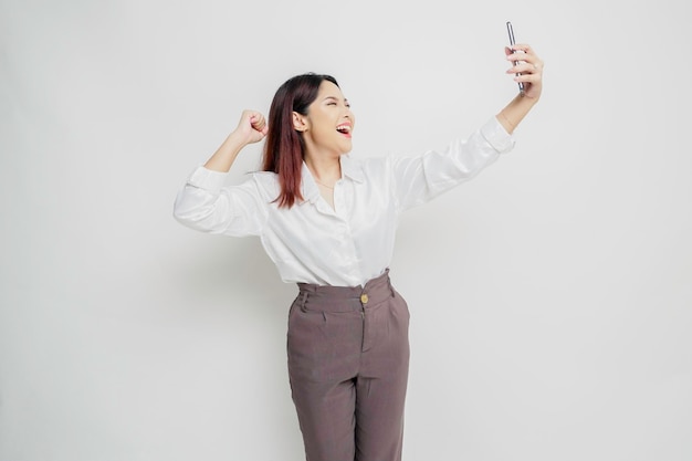 A young Asian woman with a happy successful expression wearing white shirt and holding smartphone isolated by white background