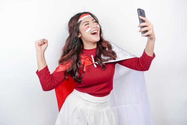 Молодая азиатская женщина с счастливым успешным выражением лица, держа в руках телефон и носящий красную головную повязку с флагом и плащ, изолированный белым фоном.