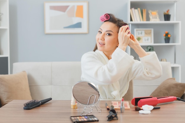 사진 긴 검은 머리를 가진 젊은 아시아 여성이 집 화장대에 앉아 아침 화장을 하며 행복하고 긍정적인 머리에 헤어 롤러를 바르고 있다