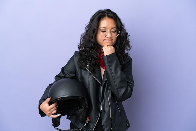Молодая азиатская женщина с мотоциклетным шлемом изолирована на фиолетовом фоне, сомневаясь
