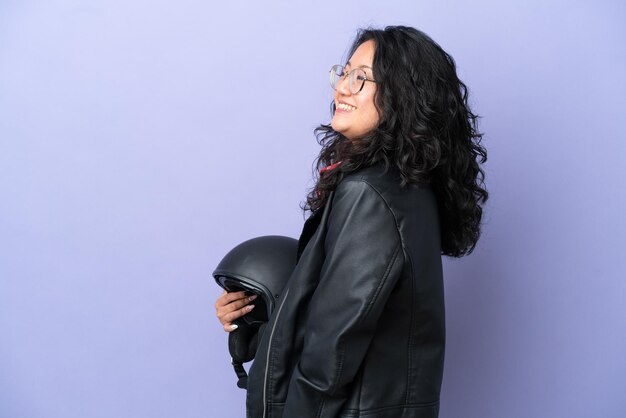 Young asian woman with a motorcycle helmet isolated on purple background laughing in lateral position