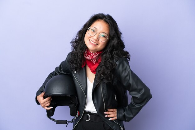 Молодая азиатская женщина с мотоциклетным шлемом изолирована на фиолетовом фоне, позирует с руками на бедре и улыбается