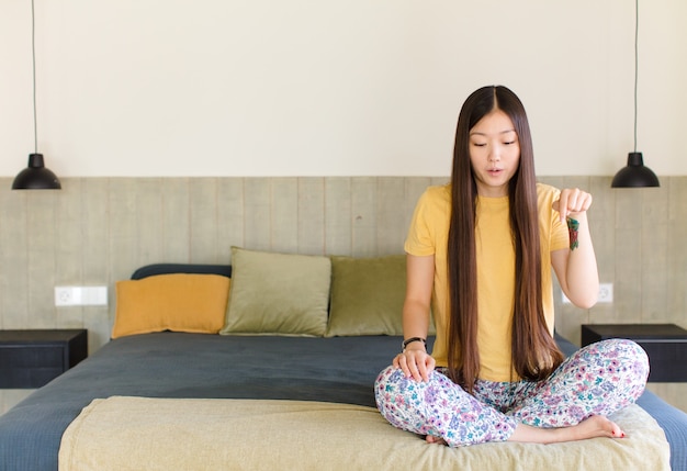 Young asian woman with open mouth pointing downwards with both hands, looking shocked, amazed and surprised