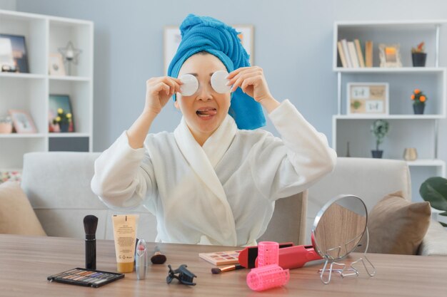 Photo young asian woman with towel on her head sitting at the dressing table at home interior holding cotton pad covering eyes with them doing morning makeup routine beauty