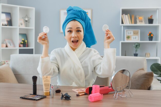 young asian woman with towel on her head sitting at the dressing table at home interior holding cotton pads happy and surprised doing morning makeup routine