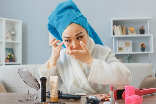 Young asian woman with towel on her head sitting at the\
dressing table at home interior looking in the mirror squeezing her\
pimples doing morning makeup routine