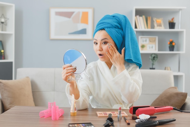 young asian woman with towel on her head sitting at the dressing table at home looking in mirror being surprised doing morning makeup routine