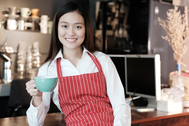 若いアジアの女性バスタブカフェカウンターの背景で笑顔でコーヒーカップを保持