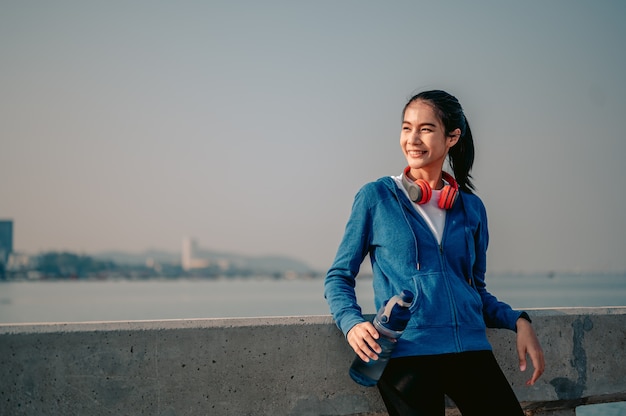 Young asian women drink water and stand to rest after jogging a morning workout in the city