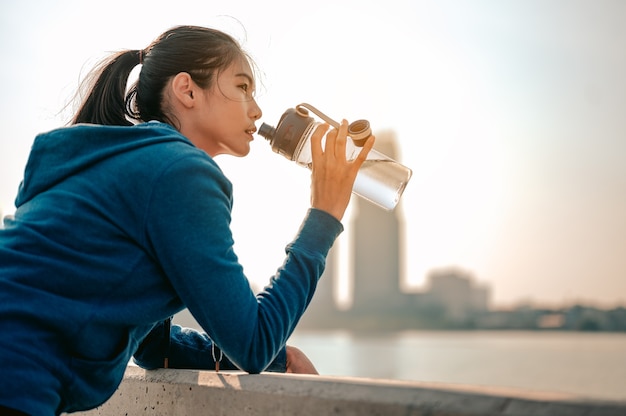 若いアジアの女性は、朝のトレーニングをジョギングした後、水を飲み、街の景色を見るために立っています