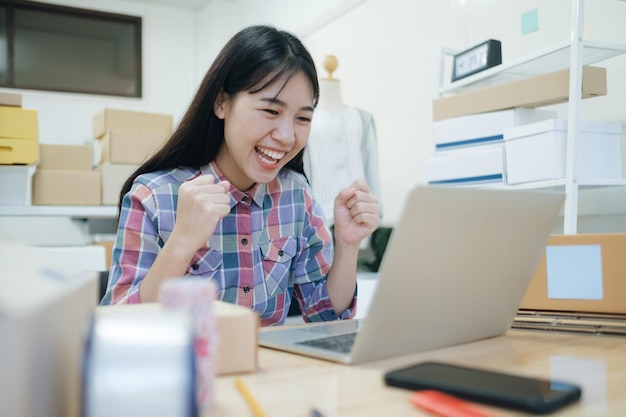 顧客からの新しい注文の後に幸せな若いアジアの女性。彼のオンラインストアの大売り出しでアジアの女性が成功したことの驚きと衝撃的な顔。オンライン販売。オンラインショッピング。