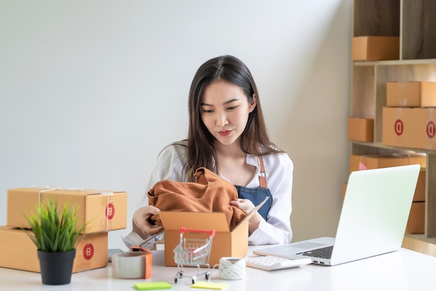 若いアジアの女性の中小企業の所有者は、自宅のラップトッププレースデスクを顧客に届けるためにオンラインで梱包しています。