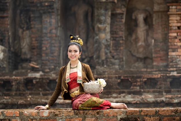 Young Asian women in Traditional dress sit on old wall  and hold silver bow ot lotus in hand.Beautiful girls in traditional costume.Thai girl in retro Thai dress.