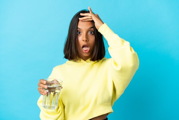 Young asiatic woman with a glass of water isolated on blue background doing surprise gesture while looking to the side