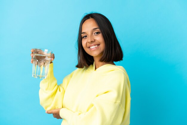 青い壁に隔離された水のガラスを持つ若いアジアの女性は、来て招待するために手を横に伸ばしています