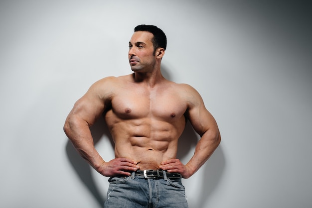 A young athlete bodybuilder poses in the studio topless in jeans near the wall. Sport.