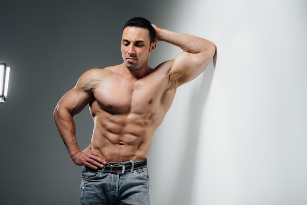 A young athlete bodybuilder poses topless in jeans near the wall. Sport.