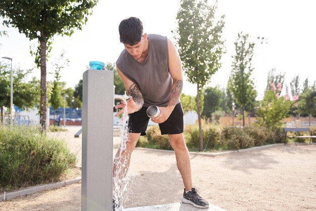 Молодой спортсмен с татуировками наполняет бутылку с водой после тренировки