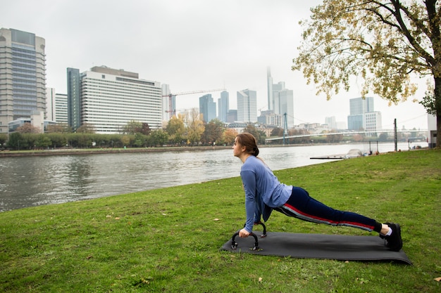Young athlete woman doing a push up with push-up bars on the\
grass, near the river, on the background of the city. sport and\
training concepts