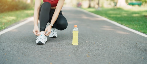 Young athlete woman tying running shoes with Energy Drink water female runner ready for jogging outside asian Fitness walking and exercise in the park morning wellness wellbeing and sport concepts