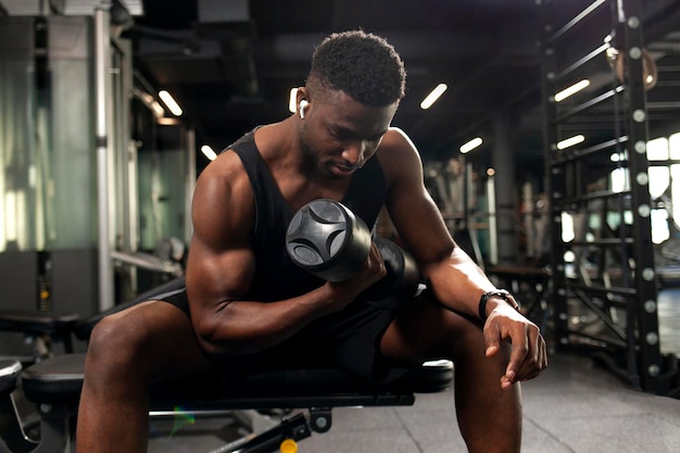 Photo young athletic african american man trains in dark gym athletic guy lifts heavy dumbbells