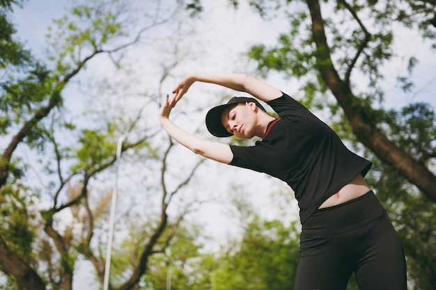 Young athletic beautiful brunette woman in black uniform, cap doing sport stretching exercises, warm-up before running or training, standing in city park outdoors