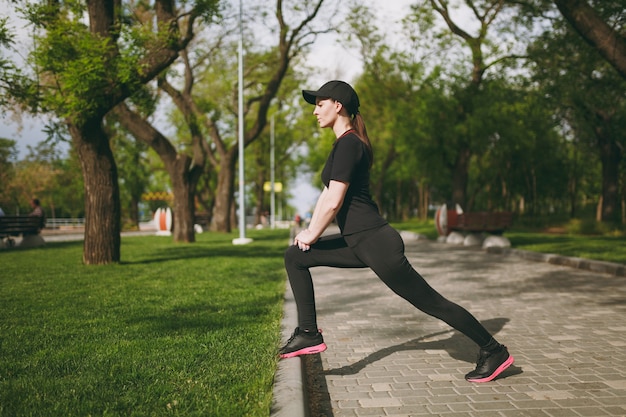 Young athletic beautiful brunette woman in black uniform, cap doing sport stretching exercises, warm-up before running or training, standing in city park outdoors