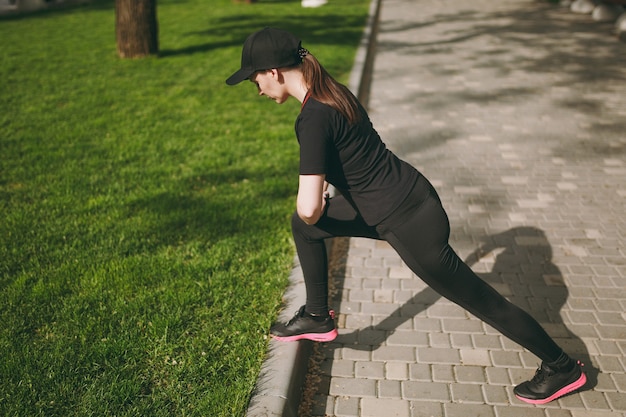 Young athletic beautiful brunette woman in black uniform, cap doing sport stretching exercises, warm-up before running or training, standing in city park outdoors