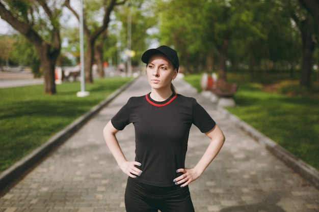 Young athletic beautiful brunette woman in black uniform and cap standing, doing sport exercises, warm-up before running, training on path in city park outdoors