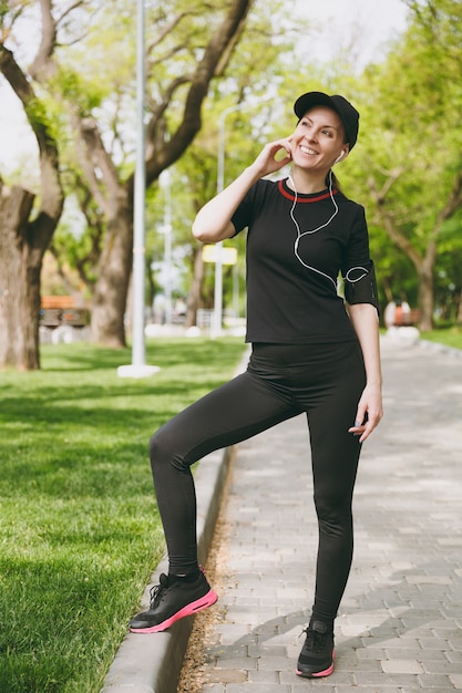 Giovane bella donna bruna atletica in uniforme nera e berretto con gli auricolari che ascolta la musica, in piedi prima di correre, allenandosi sul percorso nel parco cittadino all'aperto