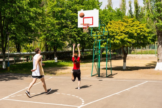 一緒にバスケットボールをする若い運動カップル-緑豊かな緑豊かな公園の屋外コートでショットを撮る女性