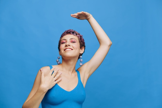 Young athletic fashion woman with colored hair and short haircut posing and dancing in blue sportswear smiling and looking at the camera on a blue monochrome background